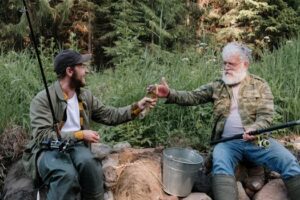 Two men share a moment while fishing in a lush, green countryside area.